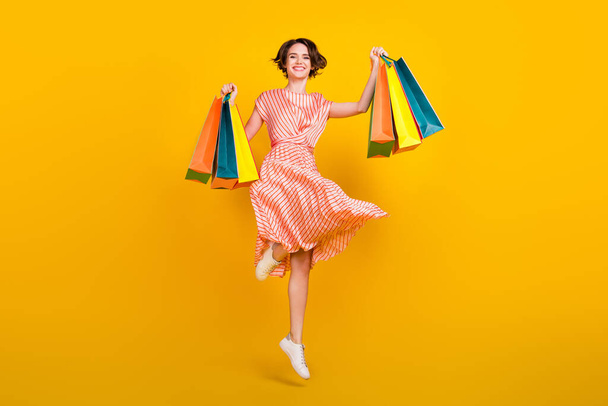 Photo of shopaholic active girl jump hold bargains wear striped dress sneakers isolated yellow color background - Fotoğraf, Görsel