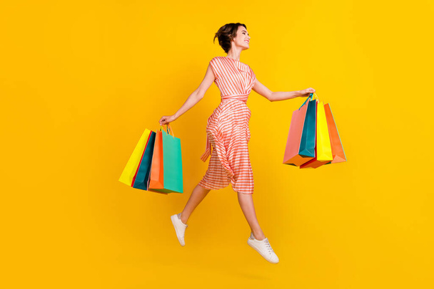 Profile photo of carefree girl jump carry store bags wear striped dress sneakers isolated yellow color background - Valokuva, kuva