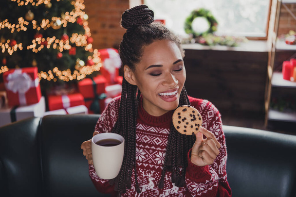 Photo of peaceful serene lady sit sofa hold coffee cup enjoy chocolate cookie wear pullover in decorated home indoors - Foto, imagen