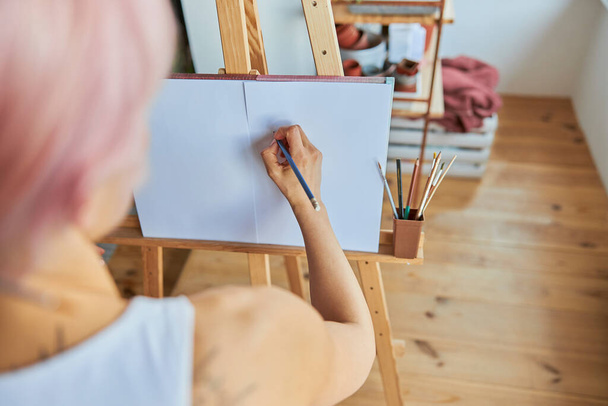 Woman painter uses graphite pencil to draw picture on blank paper at wooden easel - Fotó, kép