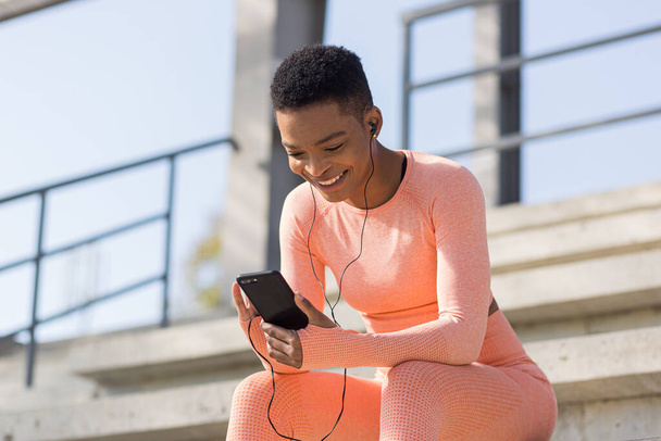 Beautiful and successful female athlete smiling listens to audio book from mobile phone during morning fitness and jogging near stadium, african american woman with mobile phone - Foto, immagini