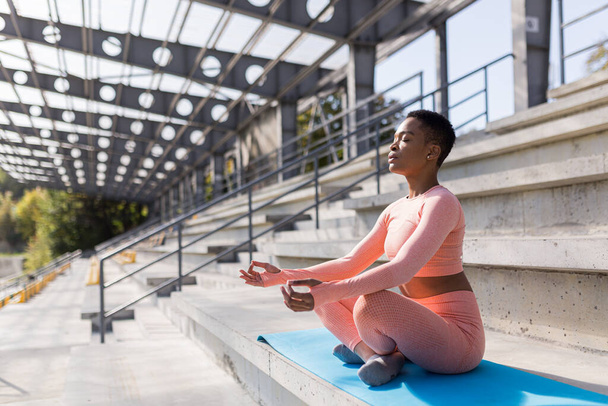 African American woman performs breathing exercises sitting on a yoga mat in the park near the stadium, resting after training - 写真・画像