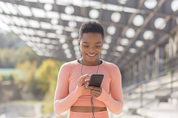 Beautiful and successful female athlete smiling listens to audio book from mobile phone during morning fitness and jogging near stadium, african american woman with mobile phone - Фото, изображение