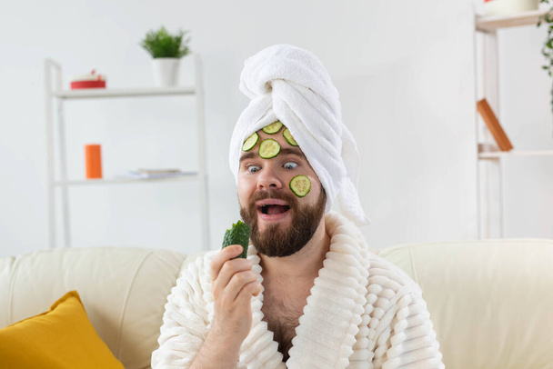 Vicces szakállas férfi, aki uborka szeleteket tart az arcán. Spa otthon, test és bőr gondozás a férfi koncepció. - Fotó, kép