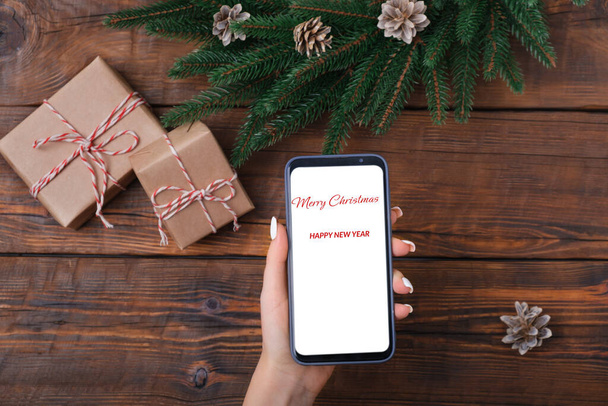 Female hands holding modern smartphone with mockup on wooden vintage table with christmas decoration. Flat lay template. Fir wreath - Photo, Image