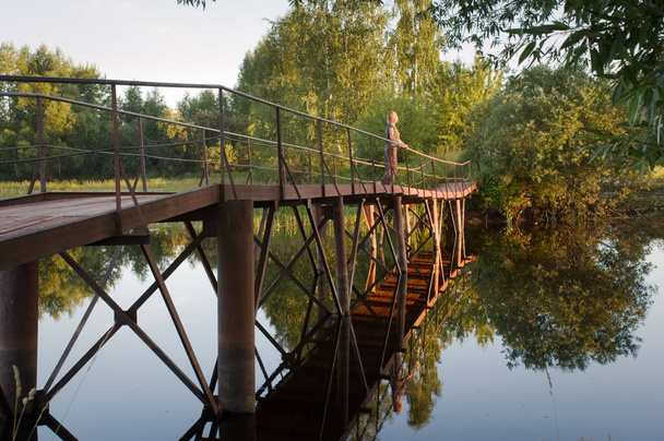 Nainen sillalla pienen joen yllä. Hiljainen vesi heijastaa siltaa, sen paaluja ja laitureita, ympäröivää luontoa. - Valokuva, kuva