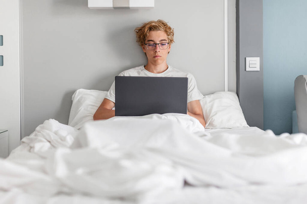 Knappe jongeman in de 20 met basic grijs t-shirt werkend op laptop pc in bed liggend met gestreepte laken kussen deken rustend tijd doorbrengend in slaapkamer thuis. Tiener jongen met laptop op campu - Foto, afbeelding