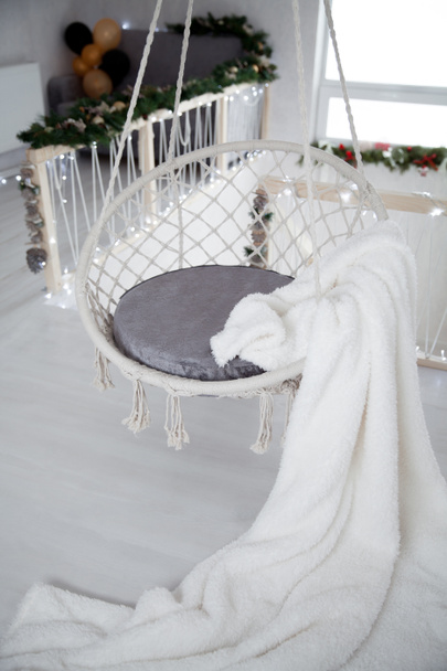 Wachten op Kerstmis. Hangende stoel met een witte bont plaid tegen de achtergrond van een kamer versierd met bloemenslingers - Foto, afbeelding
