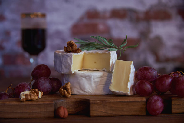 Cheese with white mold, grapes and walnuts on a wooden cutting board against the background of a brick wall. - Valokuva, kuva