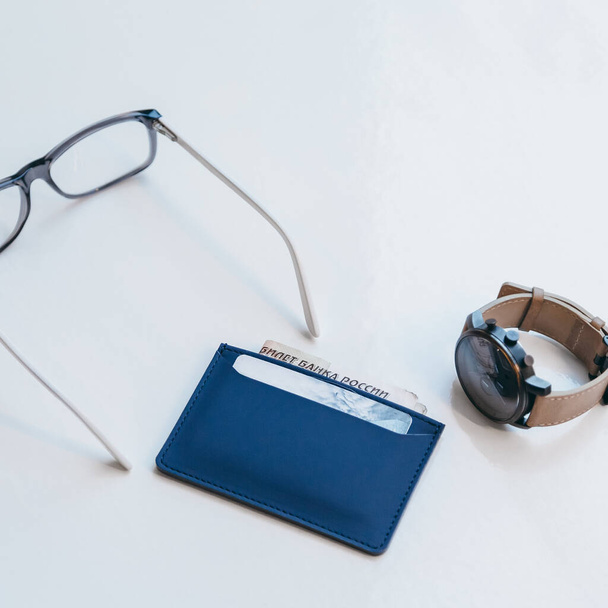 Feche a foto da bolsa fina moderna azul para cartões de crédito e notas. Acessórios minimalistas artesanais elegantes de couro ecológico. Sutura de couro liso. Perto de relógios de mão e óculos de classe.  - Foto, Imagem