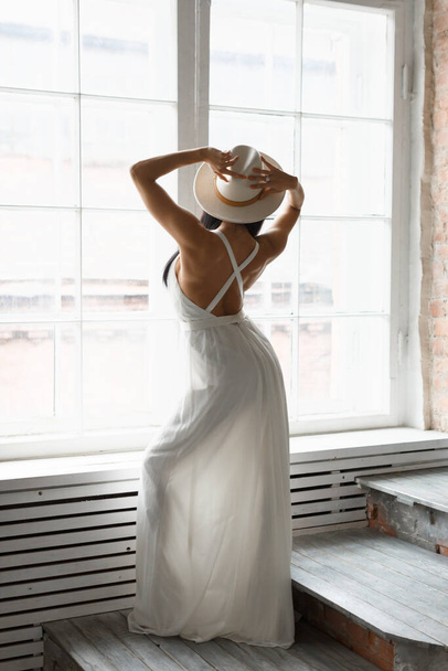 Unrecognizable slim attractive woman in white wedding chic dress stands in front of the window. Sexy bride in dress with open back. Vertical photo. Marriage concept. Waiting for the groom. - Φωτογραφία, εικόνα