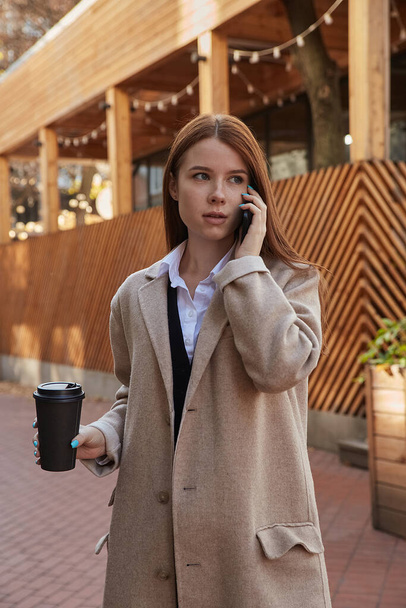 mujer caucásica en gabardina beige llamando por teléfono inteligente con taza de café - Foto, imagen