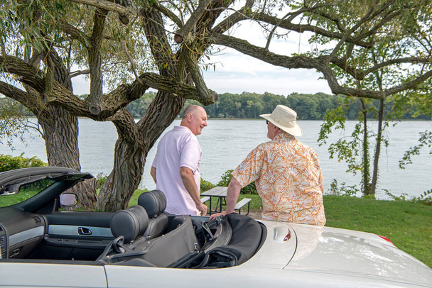 Un couple de personnes âgées LGBT mâles matures s'appuie contre leur cabriolet blanc tandis qu'ils s'arrêtent sur le bord de la route près d'une rivière avec des arbres matures et une vue panoramique. Leurs dos sont à la caméra alors qu'ils s'engagent dans la conversation. - Photo, image