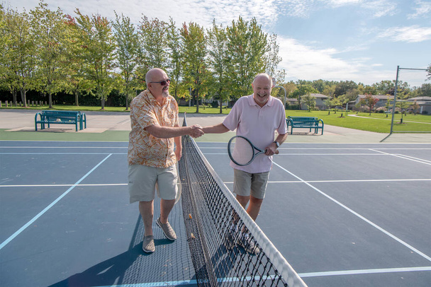 高齢の男性LGBTの結婚したカップルは、試合後にテニスコートのネット上で握手をします。夫婦は遊び心があり陽気で. - 写真・画像