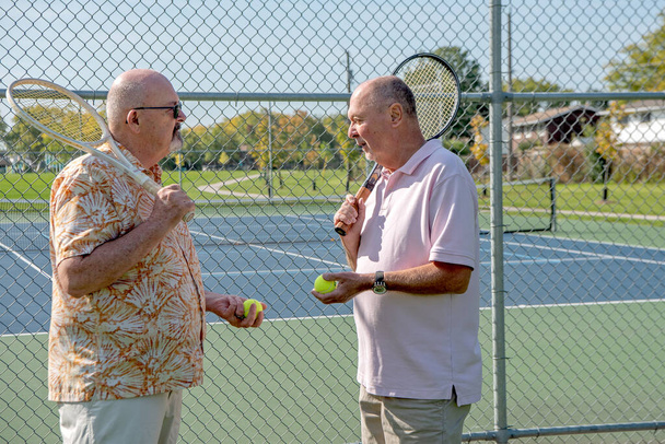A LGBT senior married couple chat while holding tennis balls and resting tennis rackets on their shoulders.  Sunny day with a public blue tennis court in the background. - Фото, изображение