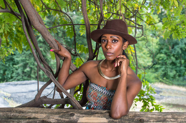 Met een bruine outback hoed, een gouden nekring en een arm die haar hoofd ondersteunt, staat een mooi zwart meisje buiten in een klein bos naar je te kijken. - Foto, afbeelding