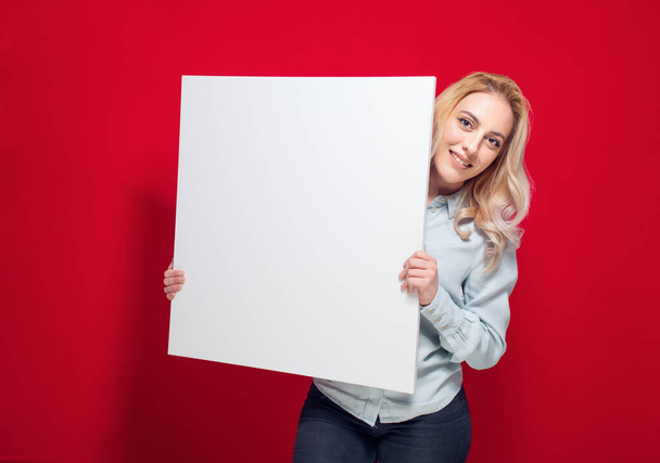 Улыбающиеся молодые женщины с белой доской, изолированные на красном фоне. Одна милая девушка показывает пустой бумажный рекламный щит с пустым местом для смс. Снимок студии - Фото, изображение