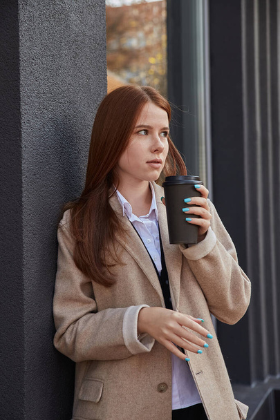 beautiful young caucasian woman in beige trench coat drinking coffee outdoors - Photo, Image