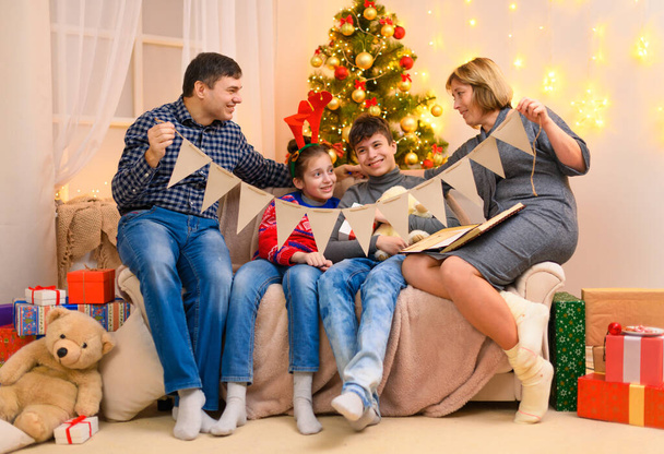 Χειμερινές διακοπές έννοια - Οικογένεια ποζάρουν στο εσωτερικό με το νέο έτος ή χριστουγεννιάτικη διακόσμηση, κάθεται σε έναν καναπέ. Παιδιά και γονείς δείχνουν εορταστική γιρλάντα από σημαίες. Φώτα και δώρα διακοπών. - Φωτογραφία, εικόνα