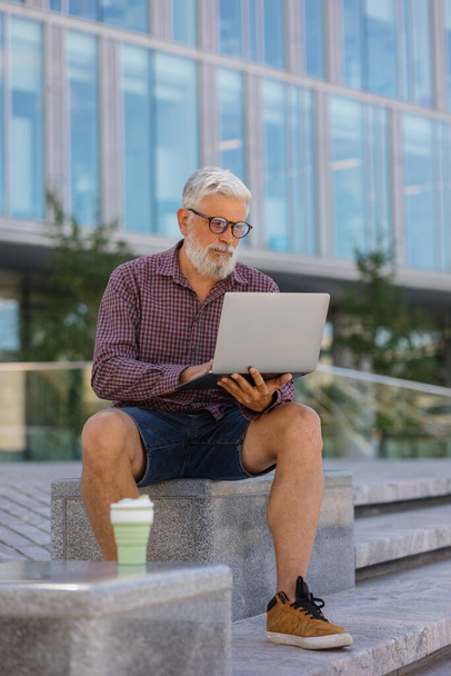 an adult gray-haired man with a beard is sitting at the office and working on a laptop - Foto, immagini