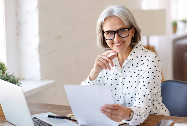 Mujer mayor sonriente sosteniendo documento financiero de papel y mirando a la computadora portátil mientras trabaja desde casa - Foto, Imagen