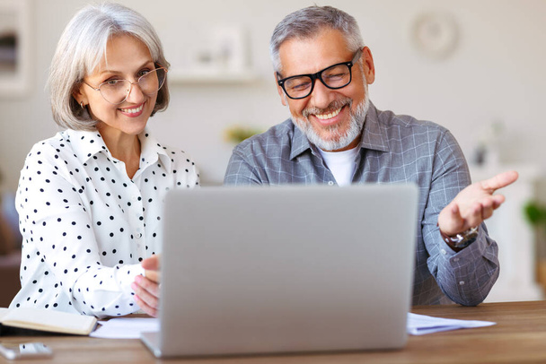 Старшая семья пара учится онлайн вместе дома, глядя на ноутбук, сидя за столом - Фото, изображение