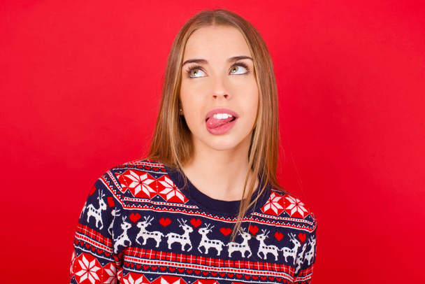νεαρό καυκάσιο κορίτσι που φοράει χριστουγεννιάτικο πουλόβερ που δείχνει γκριμάτσα να σταυρώνει τα μάτια και να δείχνει γλώσσα. Να είσαι αστείος και τρελός - Φωτογραφία, εικόνα