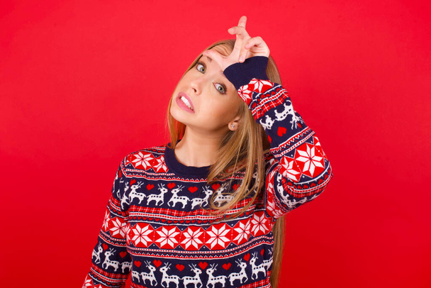 молода біла дівчина в різдвяному светрі насміхається над людьми з пальцями на лобі роблячи невдалий жест знущаючись і ображаючи
. - Фото, зображення