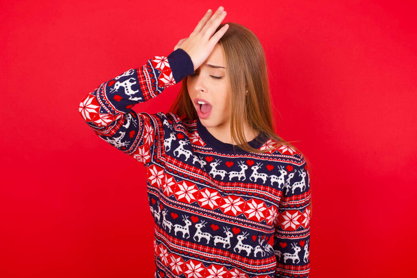 Mladá běloška na sobě vánoční svetry na červeném pozadí překvapen s rukou na hlavě pro omyl, nezapomeňte chybu. Zapomenutá, špatná koncepce paměti. - Fotografie, Obrázek
