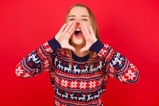 Genç beyaz kız kırmızı arka planda Noel süveteri giyiyor ve heyecanlı bir şekilde bağırıyor.. - Fotoğraf, Görsel