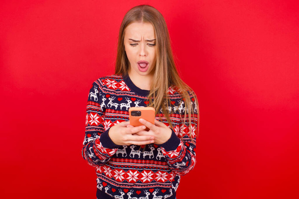 Focused Giovane ragazza caucasica che indossa maglioni di Natale su sfondo rosso usa smartphone leggere notizie sui social media o e-mail importanti - Foto, immagini