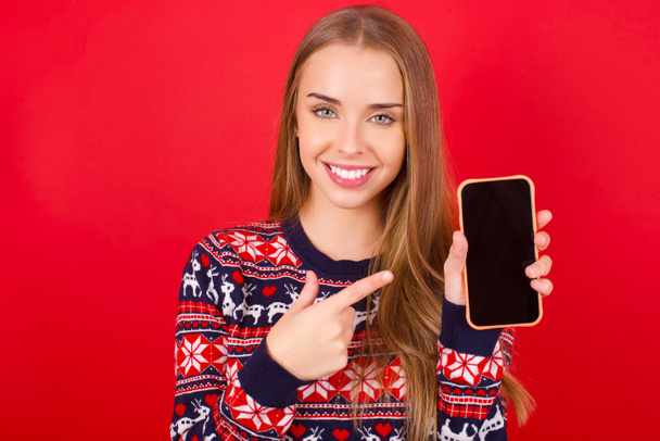 Mosolygó fiatal kaukázusi lány visel karácsonyi pulcsit piros háttér Mock up fénymásolás helyet. Mutató mutatóujj a mobiltelefon üres képernyő - Fotó, kép