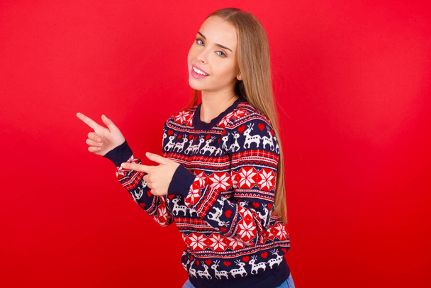 Giovane ragazza caucasica indossa maglioni di Natale sul punto di sfondo rosso a copyspace raccomandare sconti di vendita - Foto, immagini