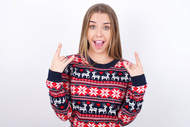 Young caucasian girl wearing christmas sweaters on white background makes rock n roll sign looks self confident and cheerful enjoys cool music at party. Body language concept. - Photo, Image
