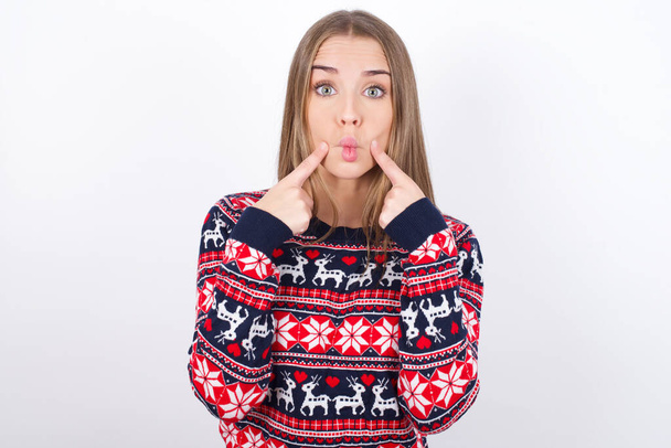 Joven chica caucásica usando suéteres de Navidad sobre fondo blanco cruza los ojos y hace que los labios de pescado divertido mueca - Foto, Imagen