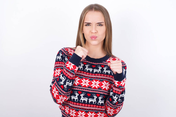 Displeased annoyed Young caucasian girl wearing christmas sweaters on white background clenches fists, gestures pissed, ready to revenge, looks with aggression at camera stands full of hate, being pressured - Foto, Bild