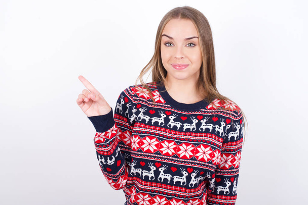 Pozitivní Mladá běloška nosí vánoční svetry na bílém pozadí se spokojeným výrazem naznačuje, v pravém horním rohu ukazuje dobrou nabídku navrhuje kliknout na odkaz - Fotografie, Obrázek