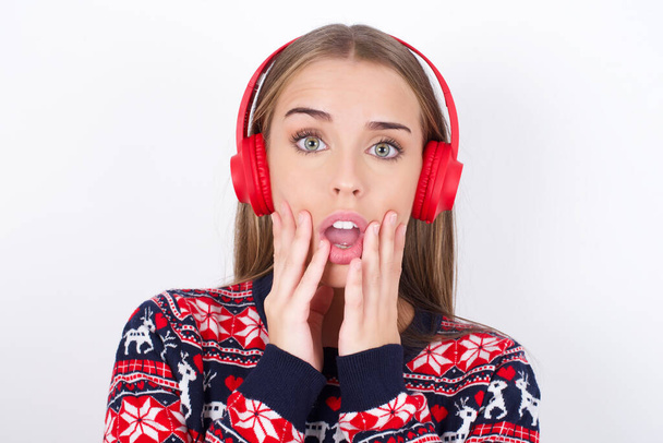 Шокированная юная кавказская девушка в рождественских свитерах на белом фоне, испуганно смотрит в камеру, держит рот широко открытым, носит беспроводные стерео наушники на ушах - Фото, изображение