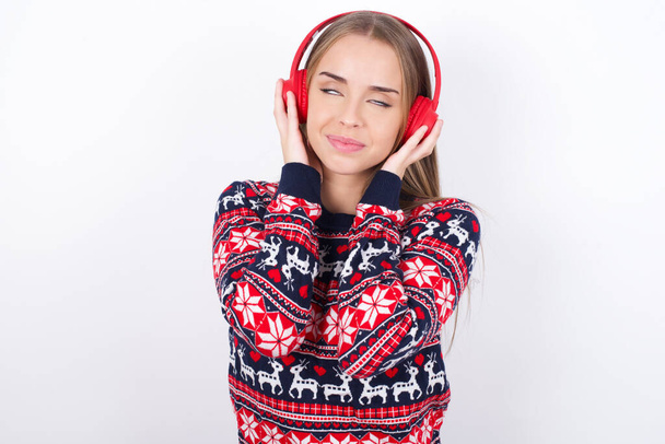 Радостная юная кавказская девушка в рождественских свитерах на белом фоне наслаждается прослушиванием приятной мелодии держит руки на стерео наушниках закрытыми глазами. Проводить свободное время с музыкой - Фото, изображение