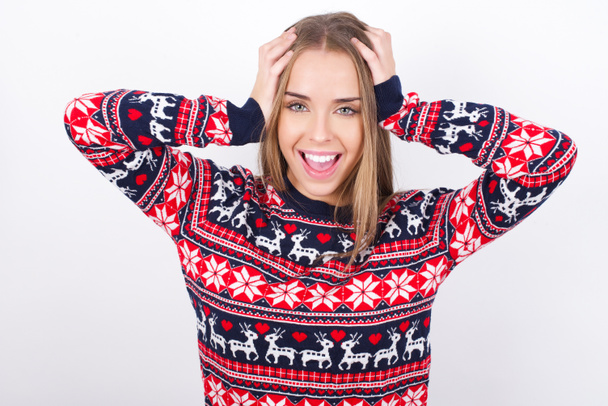 Vrolijk dolblij jong Kaukasisch meisje draagt kerst truien op witte achtergrond reageert stijgende handen over het hoofd na het ontvangen van groot nieuws. - Foto, afbeelding