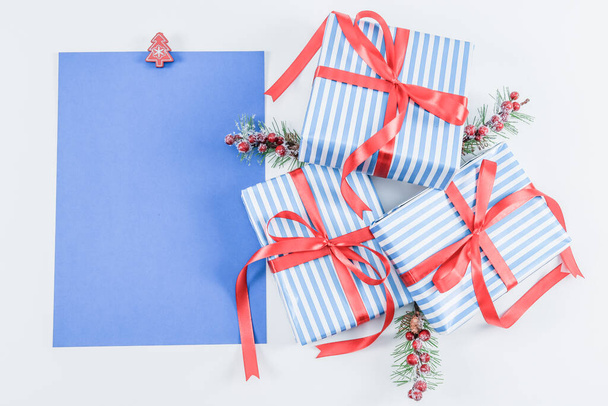 Drei Geschenkboxen in blauem Streifen mit roten Bändern, Tannenzweigen und einem leeren Blatt Papier liegen auf weißem Hintergrund, links ist in Großaufnahme Platz für Text. Weihnachts- und Neujahrskonzept. - Foto, Bild