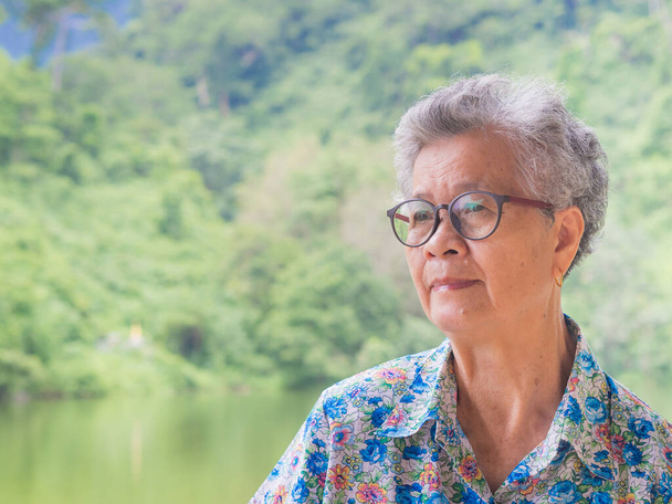 Боковой вид пожилой азиатки, улыбающейся и отворачивающейся, стоя в парке. Концепция пожилых людей и здравоохранения - Фото, изображение