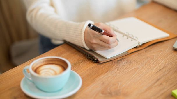 Обрезанный крупным планом образ женщины в белом свитере, пишущей или делающей заметки на блокноте, вкусный кофе на столе. - Фото, изображение