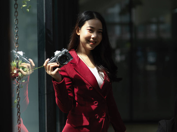 Kırmızı takım elbiseli genç Asyalı kadın. Fotoğraf retro ya da klasik fotoğraf makinesi modern stüdyo ofisinde duruyor. - Fotoğraf, Görsel