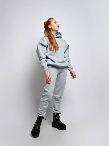 Rich slim red-haired woman in trendy grey sportswear hoodie, pants and black shoes stands sideways with eyes closed - Foto, afbeelding