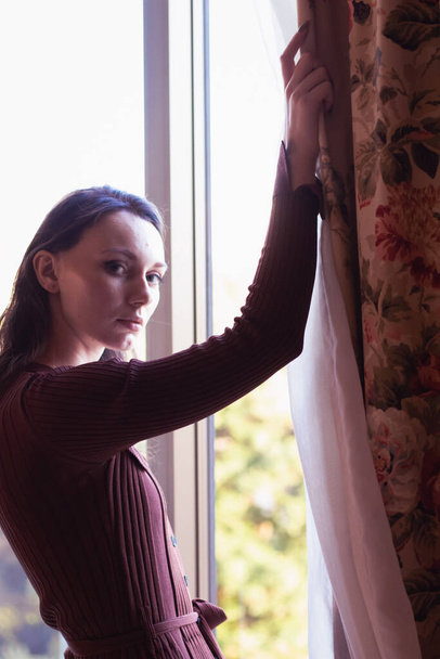 Νεαρή καυκάσια γυναίκα, στέκεται δίπλα στο παράθυρο φορώντας ένα καφέ πουλόβερ φόρεμα σε μοντέρνο στυλ δωμάτιο - Φωτογραφία, εικόνα