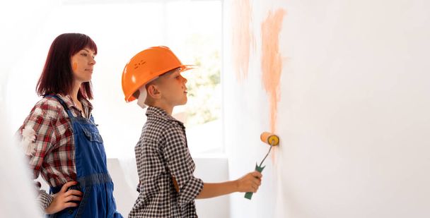 Η μαμά και ο γιος βάφουν τον τοίχο με πορτοκαλί μπογιά. έννοια ανακαίνισης σπιτιού. Ένα αγόρι με πορτοκαλί κράνος βάφει έναν τοίχο σε ένα δωμάτιο ή διαμέρισμα με ρολό. Οικογενειακή υπόθεση. banner - Φωτογραφία, εικόνα
