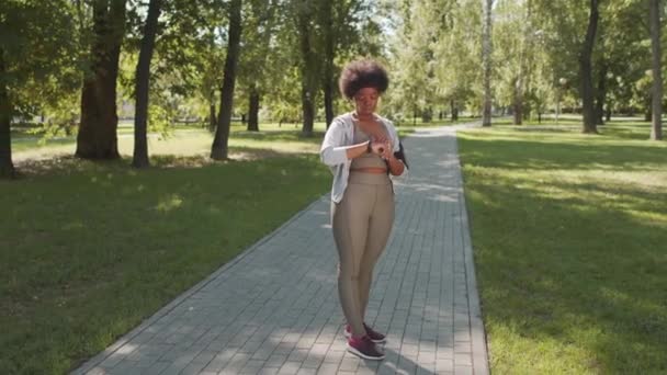 Teljes hosszúságú lassított felvétel fiatal, plusz méretű afro-amerikai nőről sportruházatban, aki időzítőt állít az okosórájára, miközben kocog a parkban a napsütéses napon. - Felvétel, videó