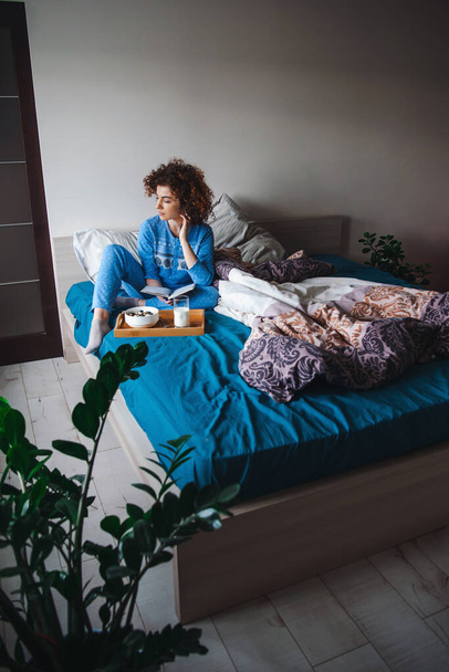Λευκή γυναίκα κάθεται με μπλε πιτζάμες στο κρεβάτι κοιτάζοντας πίσω και πέφτοντας σε σκέψεις ενώ ένα υγιεινό πρωινό την περιμένει στο κρεβάτι. Πρωινό στο κρεβάτι - Φωτογραφία, εικόνα