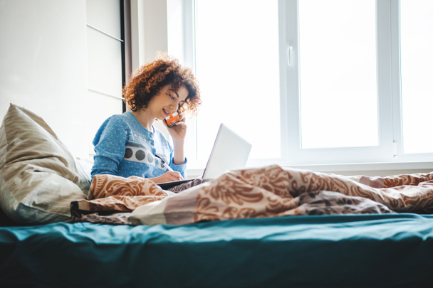 Curly woman talking on the phone straight from the bed noting homework. Online lessons - Foto, imagen